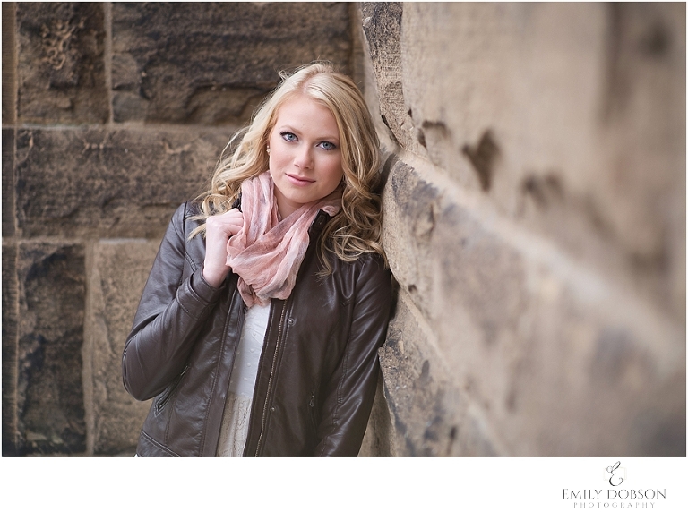 blonde HS senior leaning against a stone building in a leather jacket and scarf